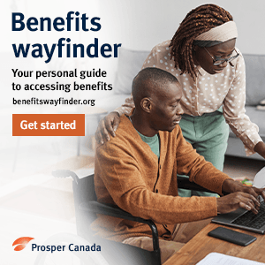 A black man in a wheel chair at a desk using a computer being helped by a black woman standing beside him.